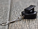 A close-up of a black retractable clip or badge retractor with a silver metal clasp, resting on a rustic gray wooden surface.
