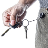 A close-up of a person's hand holding a keyring with keys, while another keyring attached to their plaid trousers has a small black circular device clipped onto the belt loop.