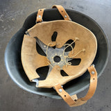 A vintage tan leather helmet interior liner with a central metal ring and adjustable straps, resting inside a dark gray helmet on a concrete floor.