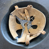 A vintage tan leather helmet interior with adjustable straps and a clear plastic liner, resting inside a black helmet on a dark concrete floor.