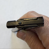 A close-up of a hand holding a Colt 40 Cal 220mm bullet fragment trace cartridge in a dark metallic finish, positioned over a white sheet of paper on a gray floor.