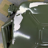 An olive green metal fuel can with peeling white paint, shown on a patterned gray carpet with a person's shoe visible in the top right corner.