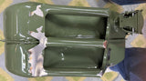 An olive green metal automotive part, likely a fender or bumper component with a cutout, lying on a patterned gray and yellow floor. A person's legs and shoes are visible in the top right corner.