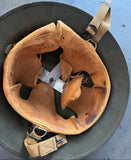 A vintage tan leather helmet interior lining resting inside a dark olive green metal helmet, with tan webbing straps and metal buckles visible.