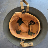 An olive green military-style helmet with a tan leather interior lining and tan webbing straps, photographed from above on a gray concrete floor.