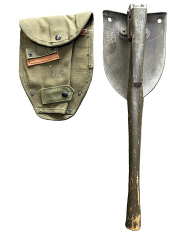 A vintage military-style shovel with a dark metal blade and wooden handle, shown next to its olive green canvas sheath on a textured gray concrete surface.