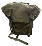 An olive green canvas military-style backpack with a roll-top closure, adjustable straps, and metal rivets, photographed on a concrete floor.