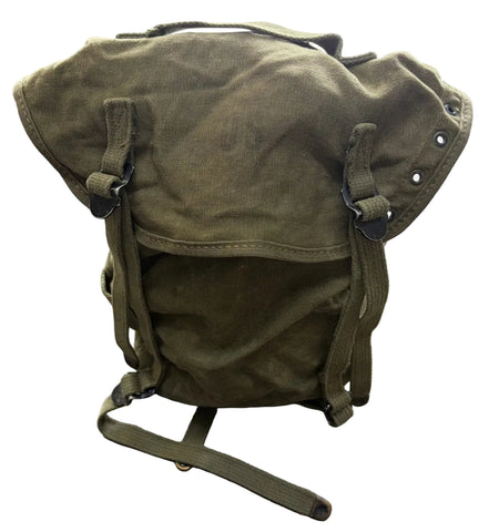 An olive green canvas military-style backpack with a roll-top closure, adjustable straps, and metal rivets, photographed on a concrete floor.