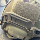 Close-up of a tactical helmet cover in a camouflage pattern with a tan plastic attachment bracket and a hook-and-loop patch.
