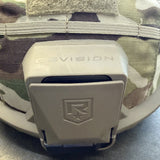 Close-up of a tan Revision tactical helmet accessory mounted on a camouflage helmet, with the brand name REVISION debossed on the front.