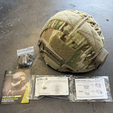 A camouflage tactical helmet cover with beige fleece lining and hook-and-loop patches, displayed on a concrete floor with included accessories and paperwork: a booklet, small hardware bag, and two instruction sheets.