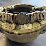 Close-up of a tactical helmet or headgear with a camouflage exterior and a tan quick-release strap and buckle closure. The center buckle has an embossed logo.