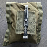 An olive green tactical pouch with a front hook-and-loop patch and a central metal clip, laid flat on a gray concrete surface.