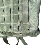 Close-up of an olive green tactical backpack or gear bag showing MOLLE webbing and heavy dirt smudging on the fabric, resting on a gray concrete surface.