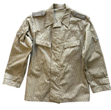 A military-style field jacket laid flat on a textured gray concrete floor. The jacket features a vertical striped pattern in olive drab and tan, with two chest flap pockets, shoulder epaulets, and buttoned cuffs.