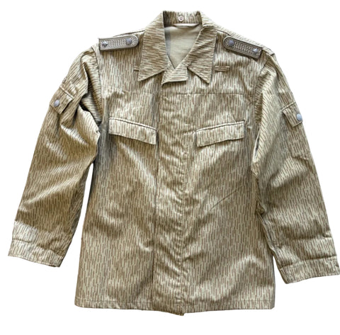 A military-style field jacket laid flat on a textured gray concrete floor. The jacket features a vertical striped pattern in olive drab and tan, with two chest flap pockets, shoulder epaulets, and buttoned cuffs.