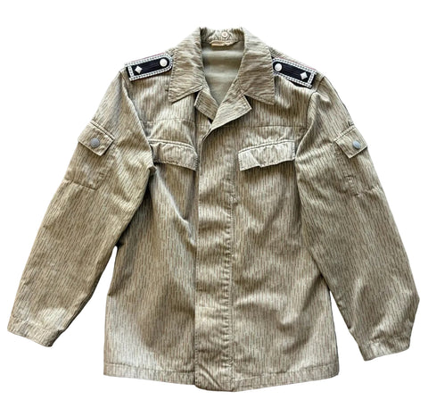 A military-style field jacket in a beige and brown vertical stripe pattern, laid flat on a gray concrete floor. The jacket features a pointed collar, shoulder epaulets with silver buttons, a button-front closure, and multiple flap pockets.