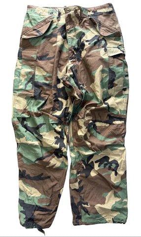 A pair of vintage woodland camouflage cargo pants laid flat on a gray concrete floor. The pants feature a classic four-color camo pattern of green, brown, black, and tan, with a drawstring waist and multiple utility pockets.