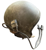 A vintage olive green military helmet with its metal chin strap and buckle attached, resting on a concrete floor.