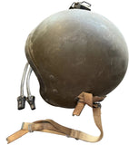 A vintage military-style helmet in olive drab green with a tan chin strap, shown on a gray concrete floor next to its black cables.