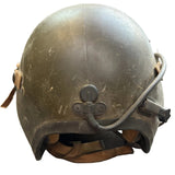 A vintage olive drab military helmet with a wired communication system and tan chin straps, resting on a concrete floor.