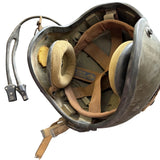 A vintage military-style helmet with a dark gray metal shell, tan leather straps, and beige foam ear cushions, shown on a concrete floor.
