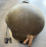 A vintage military-style helmet in olive drab green with a tan chin strap, resting on a gray concrete floor next to black cables with connectors.