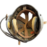 A vintage military-style helmet with a metal shell, tan interior padding, and foam ear muffs, photographed on a concrete floor.