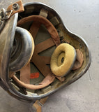 A vintage military-style helmet with a dark gray metal shell, tan interior padding, and a yellow foam ear cup, photographed on a concrete floor.