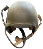 A vintage military-style helmet in olive drab green with tan straps and a black headset microphone, resting on a gray concrete floor.