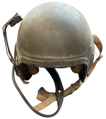 A vintage military-style helmet in olive drab green with tan straps and a black headset microphone, resting on a gray concrete floor.