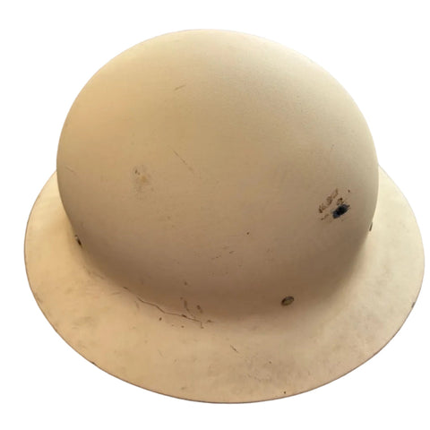 A vintage cream-colored hard hat with a wide brim, resting on a dark gray concrete floor. The hard hat shows some scuffs, dirt, and minor damage, including a small dark spot on the right side.