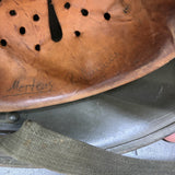 A close-up of a vintage brown leather helmet visor with circular ventilation holes, featuring handwritten names 'Mertens' and 'Robinson' on the inside, resting on a gray helmet shell.