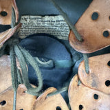 A close-up of a vintage brown leather shoebox lid with circular cutouts, showing a paper label with German text and dark green laces.